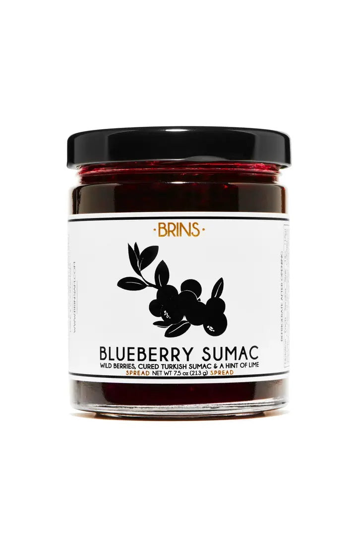 A glass jar of BRINS Wild Blueberry Sumac Spread and Preserve features a white label with a black berry illustration and detailed ingredient and net weight information. Crafted by BRINS using wild blueberries and vibrant Turkish sumac, this preserve showcases distinctive flavors elevated by single origin spices.