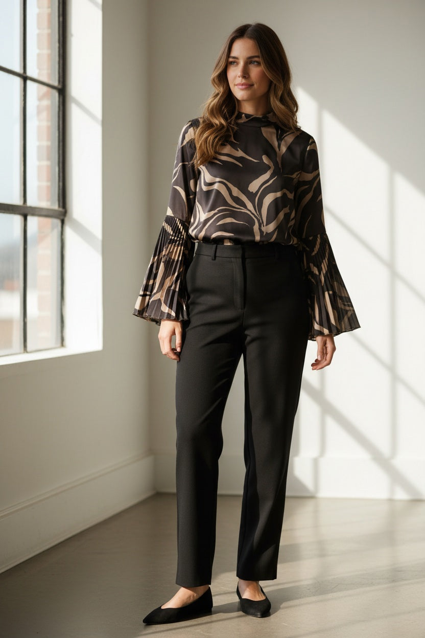 A woman poses against a brown background wearing the MILVA MI black and gold flare sleeve blouse by MILVA, paired with high-waisted black pants. Her dark hair is styled in a low bun.