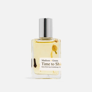 A clear glass bottle with a silver cap holds yellow, fast-absorbing aromatherapy oil infused with visible herbs and eucalyptus. The label reads “MADISON GREEN - TIME TO SHINE CONFIDENCE BOOST AROMATHERAPY BODY OIL MINI.”.