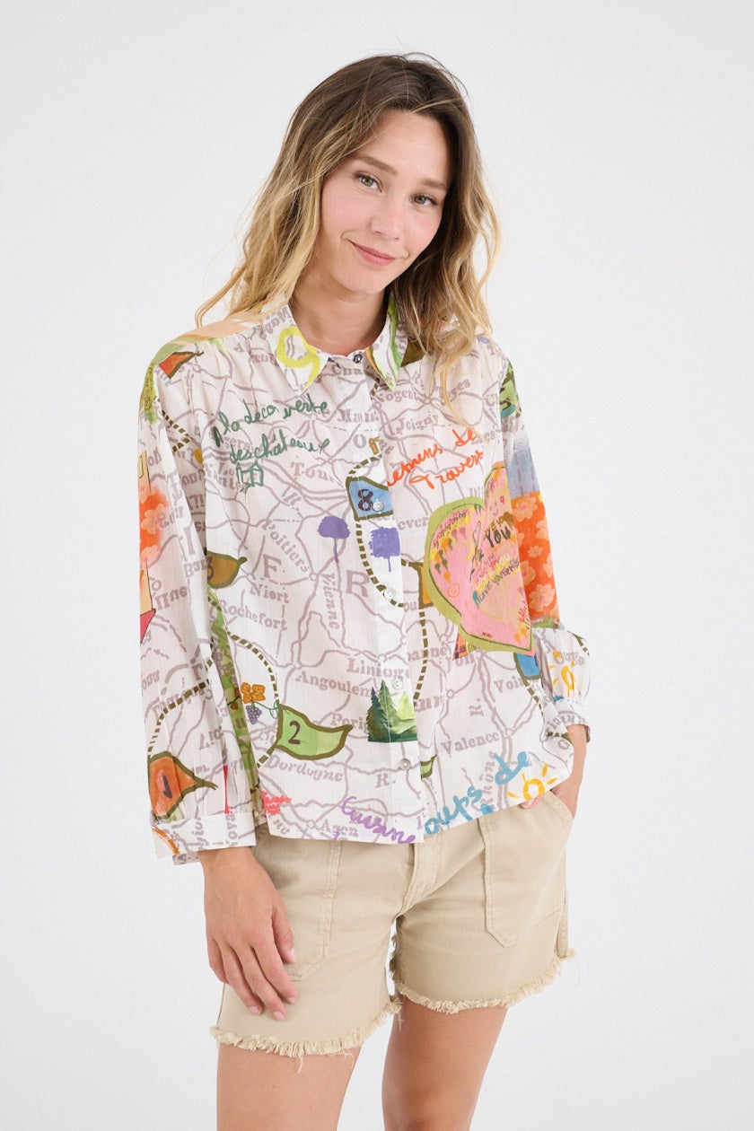 A smiling woman with long wavy hair wears the STORIATIPIC - SUZIE GABIE BLOUSE, a colorful off-the-shoulder cotton top with map and floral patterns, paired with beige frayed shorts against a plain white background.
