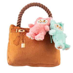A French Bulldog sits indoors in a bright, cozy room, holding the HAUTE DIGGITY DOG LABOWWOW HANDBAG DOG TOY—a durable plush handbag adorned with pink and blue teddy bear keychains—in its mouth.