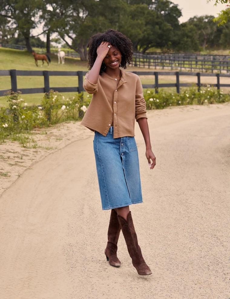 A person wearing a tan shirt, the FRANK AND EILEEN - SHANNON CLASSIC DENIM SKIRT IN 1978 WASH by FRANK & EILEEN, and brown cowboy boots stands on a dirt road.