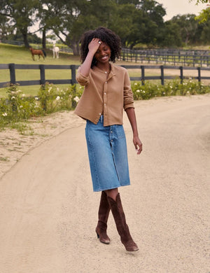 A person wearing a tan shirt, the FRANK AND EILEEN - SHANNON CLASSIC DENIM SKIRT IN 1978 WASH by FRANK & EILEEN, and brown cowboy boots stands on a dirt road.