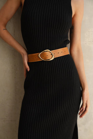 A person wearing a sleeveless, ribbed black dress with a tan belt featuring a unique, rounded buckle stands against a light-colored wall, with one hand resting on their hip.
