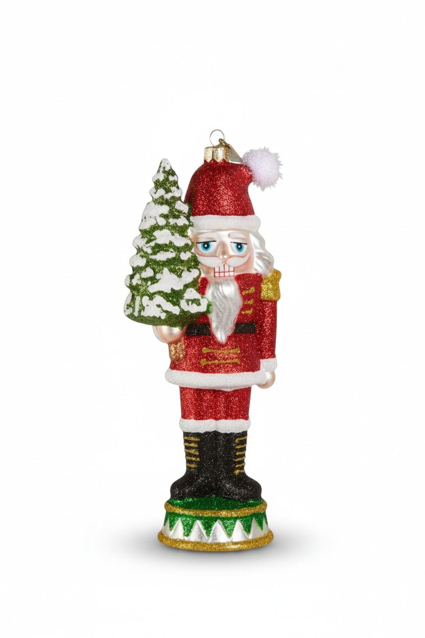 A GLITTER POPCORN NUTCRACKER ORNAMENT made of glass and resin, featuring a glittery nutcracker dressed as Santa Claus and holding a snow-dusted evergreen tree.