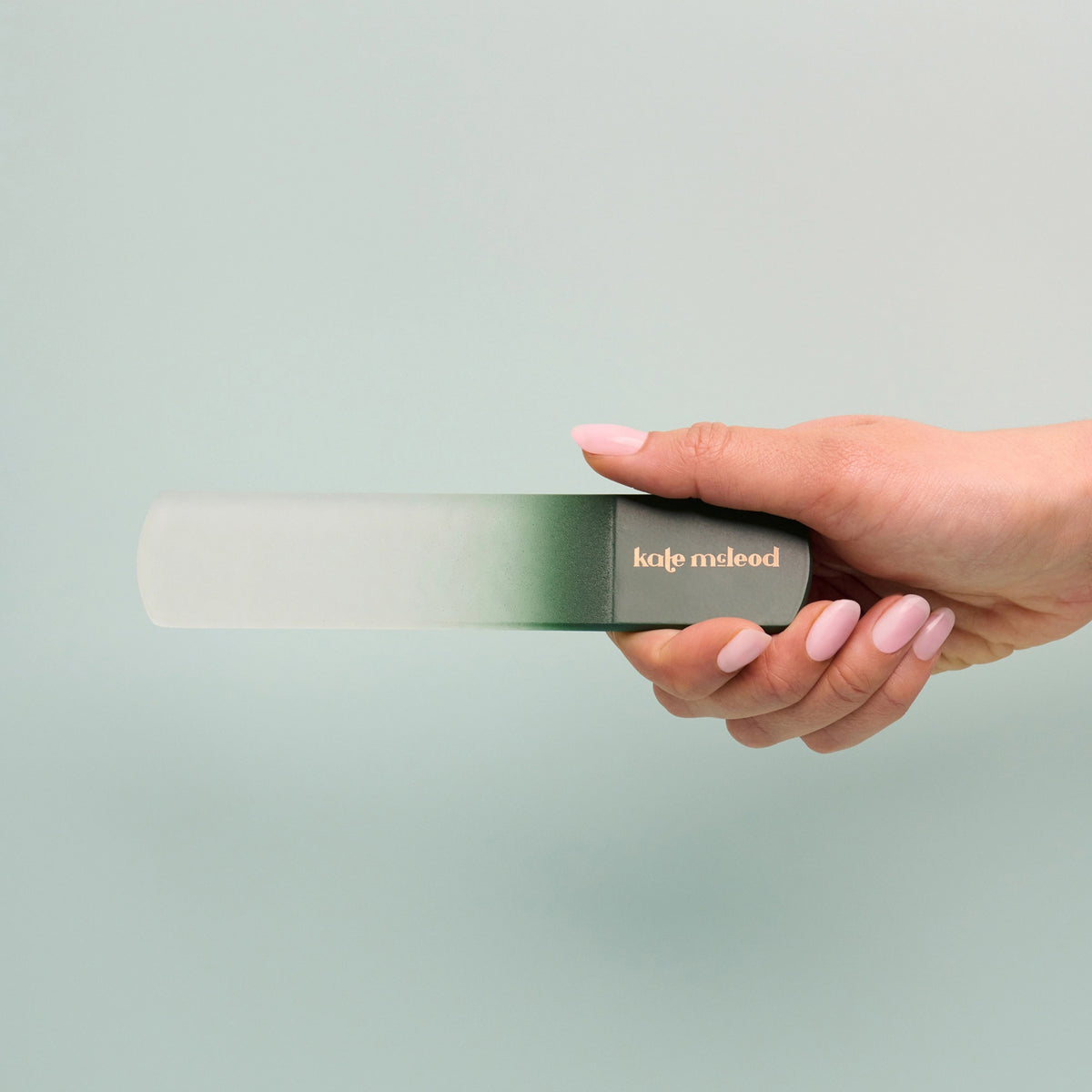 A hand with pale pink nails holds a cylindrical, green-gradient object labeled "KATE MCLEOD," designed for callus care—the light blue-green background enhances its minimalist aesthetic.