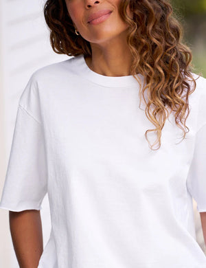 A person with wavy brown hair stands outdoors, partially smiling against a blurred background, wearing the FRANK & EILEEN PERRY NYC Crewneck Tee in Vintage Jersey White.