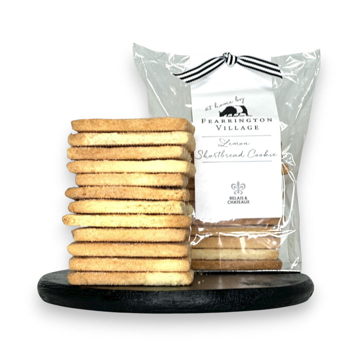 A stack of buttery, crisp FEARRINGTON SIGNATURE COLLECTION - HOUSEMADE LEMON SHORTBREAD COOKIES stands on a black round surface. Behind the stack, a clear plastic bag containing more cookies is tied with a black and white ribbon and features a label reading &quot;FEARRINGTON HOUSE Lemon Shortbread Cookie&quot; with decorative details from The Fearrington House Restaurant.