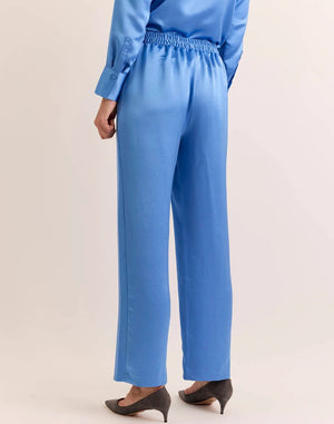 Model wears the HINSON WU - HOSTESS LIQUID SATIN PANT IN SKY BLUE by HINSON WU, featuring a relaxed straight leg and elastic waistband in luminous, light-catching sky blue satin. Styled with the matching long-sleeve top, they are pictured from the back against a neutral backdrop and complete the look with gray pointed-toe shoes.