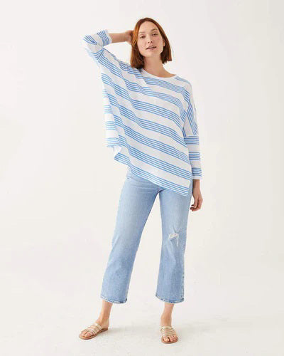 A woman with wavy brown hair models the MERSEA - CATALINA SLUB TEE—an oversized, long-sleeved white top with thin gray stripes—by MERSEA. She pairs it with blue jeans and stands against a plain, light background, hands behind her back.