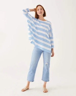 A woman with wavy brown hair models the MERSEA - CATALINA SLUB TEE—an oversized, long-sleeved white top with thin gray stripes—by MERSEA. She pairs it with blue jeans and stands against a plain, light background, hands behind her back.