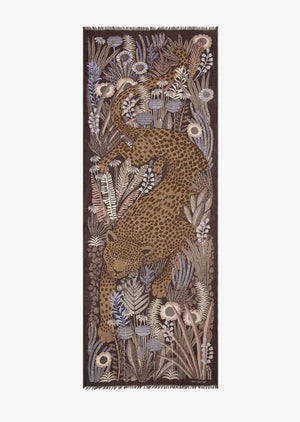 A person with shoulder-length gray hair stands facing the camera, wearing a white shirt and blue jeans. Adorning their neck is an INOUI EDITIONS - ROUSSEAU WOOL SCARF by INOUI, featuring an artisan-made brown fabric with a floral and animal pattern. The background is plain white.