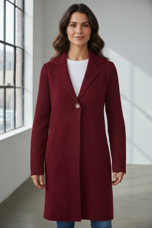 A woman stands indoors wearing the KINROSS CASHMERE Rib Sleeve Notch Collar Long Cardigan in Cordovan over a white top and blue jeans. This timeless piece from KINROSS features classic ribbed sleeves, combining elegant style with cozy practicality for cold-weather layering. With wavy brown hair and a slight smile, she stands near large windows bathed in natural light.