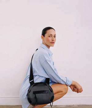 A person in a light beige suit carries the DANIELLA LEHAVI - BONNIE TRAVEL CROSSBODY, a large brown vegan bag with black zipper and strap, shown from shoulders to mid-thigh against a plain background.