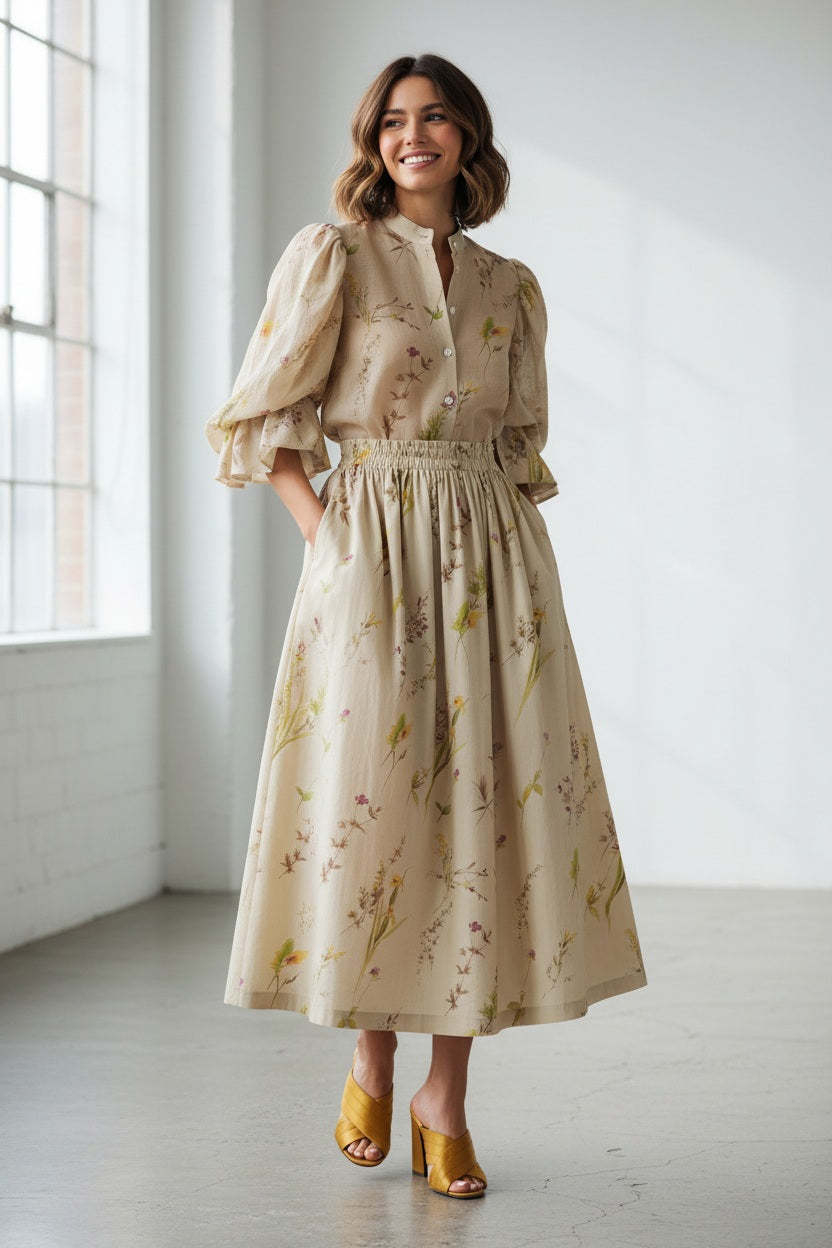 A woman stands against a plain background wearing the MIRTO 1956 Beige Floral Print Midi Skirt, paired with puffed three-quarter sleeves, a wide tan belt, and brown open-toe heels. Her hair is styled in a high bun with a decorative accessory.