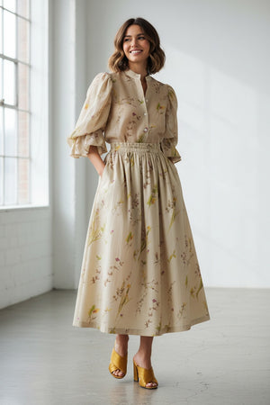 A woman stands against a plain background wearing the MIRTO 1956 Beige Floral Print Midi Skirt, paired with puffed three-quarter sleeves, a wide tan belt, and brown open-toe heels. Her hair is styled in a high bun with a decorative accessory.