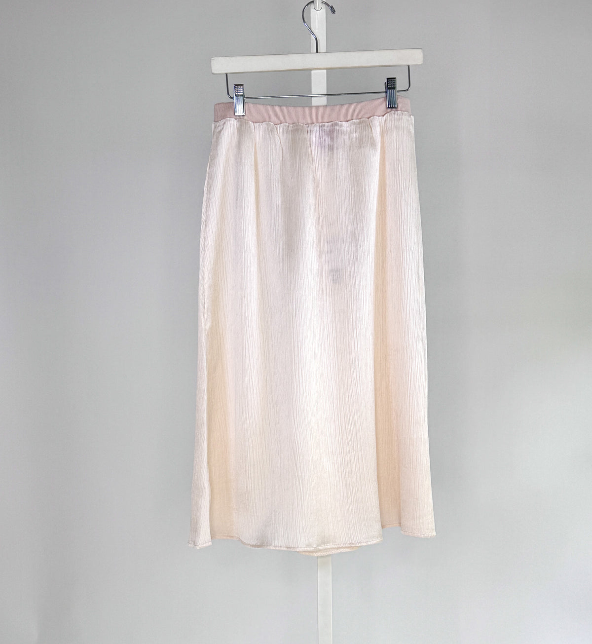 A woman in a sleeveless black dress with a wide belt stands against a plain backdrop, holding a large black handbag. She wears black shoes and layers the ROSE AND CROWN - SATIN RIBBED SKIRT by ROSE &amp; CROWN under her dress.