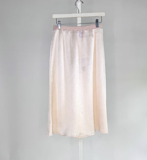 A woman in a sleeveless black dress with a wide belt stands against a plain backdrop, holding a large black handbag. She wears black shoes and layers the ROSE AND CROWN - SATIN RIBBED SKIRT by ROSE & CROWN under her dress.