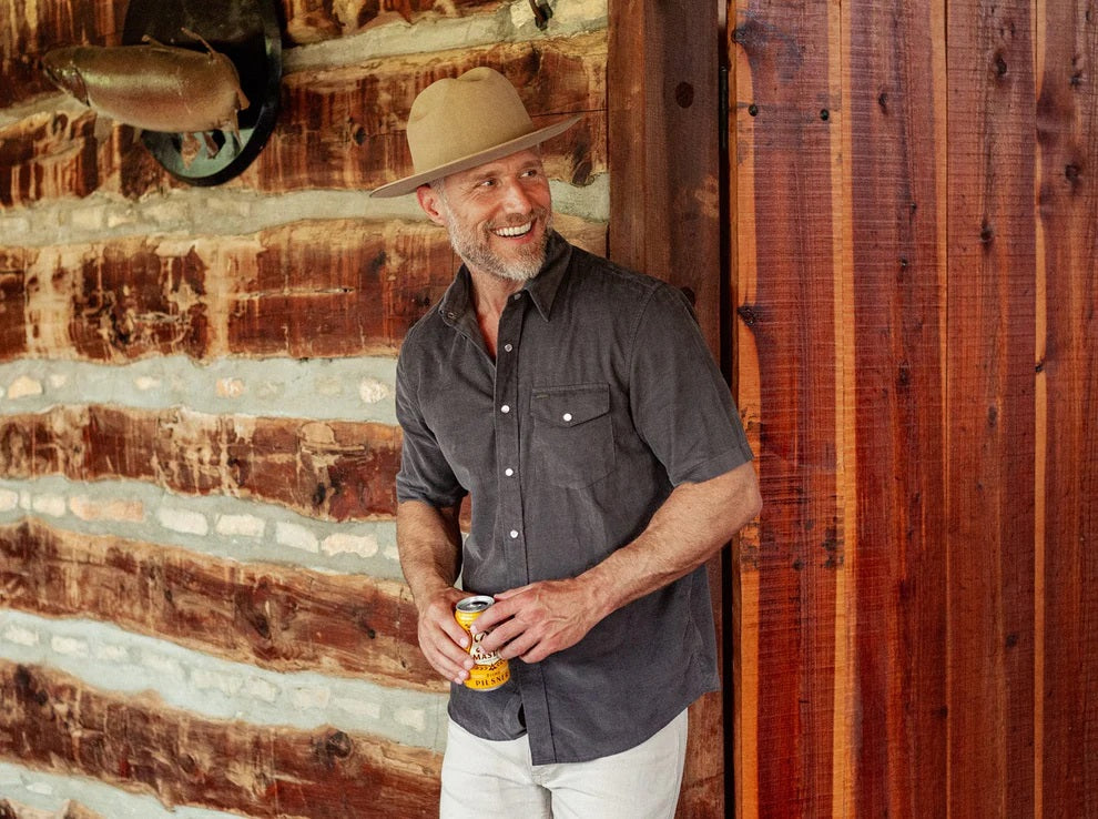 A smiling man in a brown hat and the CRIQUET Short Sleeve Corduroy Pearl Snap Shirt in Iron Horse holds a canned drink in his right hand, standing against a white background.