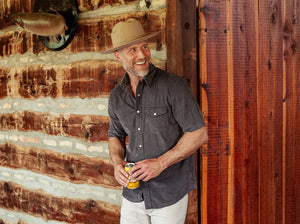 A smiling man in a brown hat and the CRIQUET Short Sleeve Corduroy Pearl Snap Shirt in Iron Horse holds a canned drink in his right hand, standing against a white background.