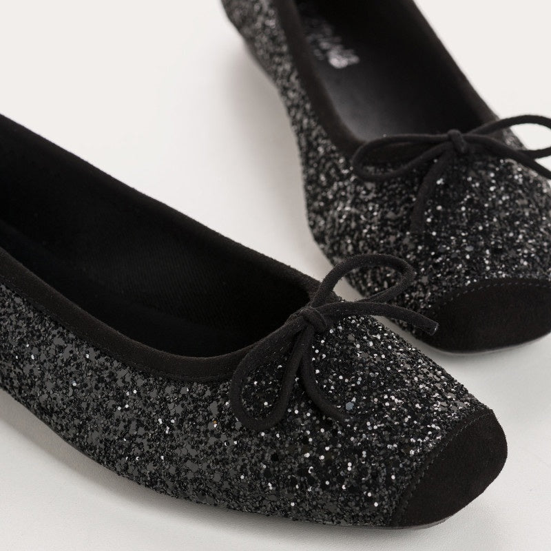 The REQINS Harmony Sequin Ballerina Flats in Noir feature black sequins, black toe caps, and small bows—bringing a touch of everyday luxury against a plain light background.