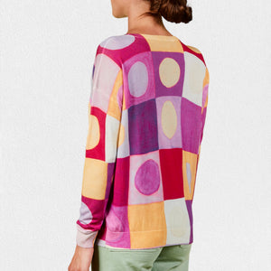 A person wearing the IN BED WITH YOU - BATWING SLEEVE V NECK JUMPER stands against a plain, light-colored background. The colorful geometric pattern featuring squares and circles in shades of pink, purple, red, yellow, and white adds flair to this stylish piece by the Italian brand IN BED WITH YOU/CLAUDIOLAI SRL. The person is visible from the shoulders down to the waist.