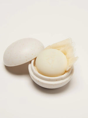 Two round soaps are in open speckled white KATE MCLEOD - THE MINI TRAVEL CASE. The larger soap, similar to a Mini Body Stone, is light beige and wrapped in cloth; the smaller one is cream-colored. Both cases have detached round lids on a flat white surface.
