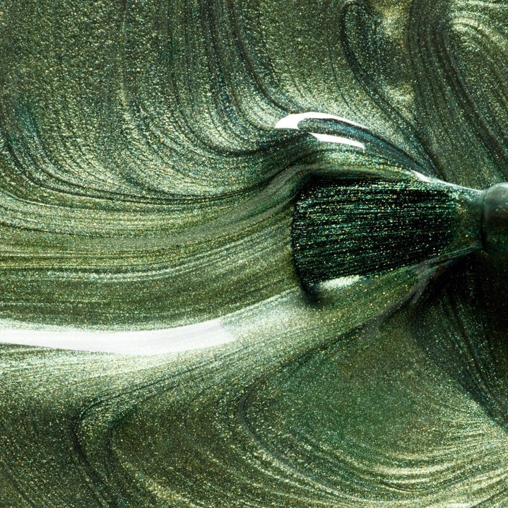 A hand with metallic green nails holds a bottle of MANUCURIST - GREEN™ NAIL POLISH IN GROOVE from MANUCURIST INC, perfectly matching the light green shirt in the background.
