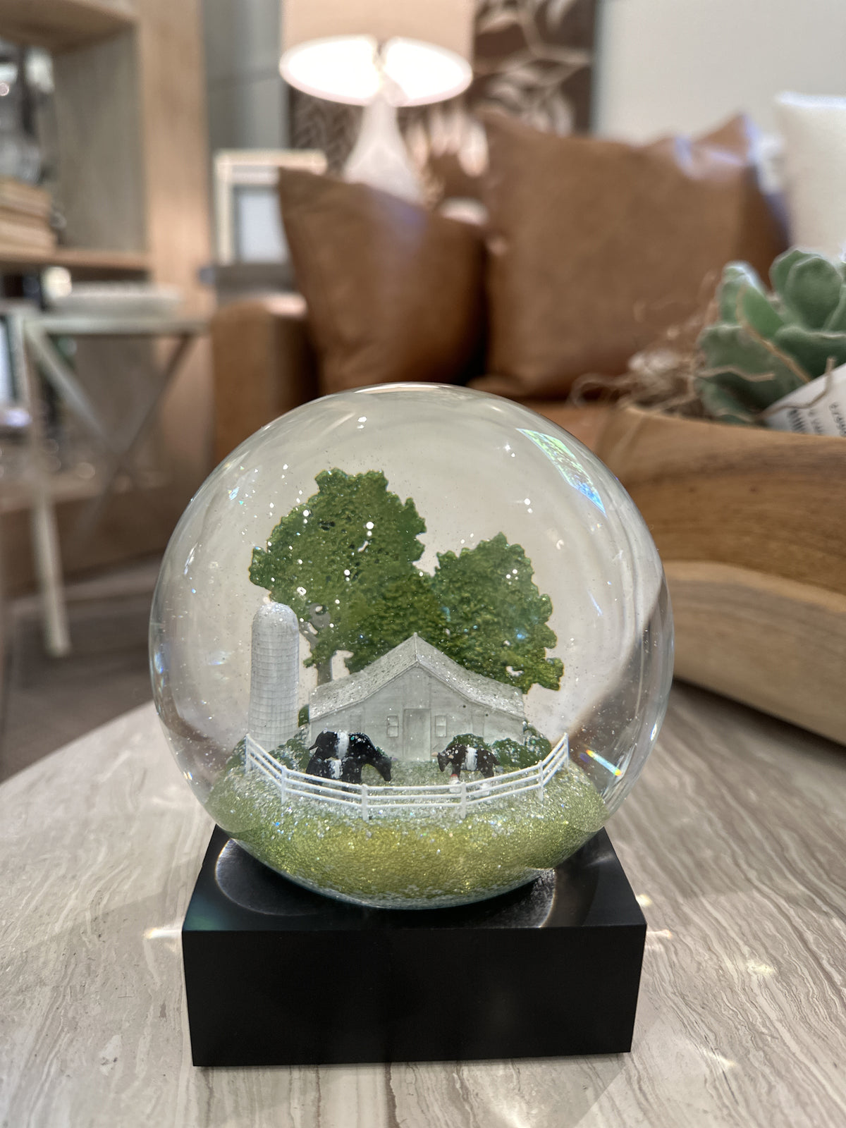 The FEARRINGTON SIGNATURE COLLECTION - FEARRINGTON BELTIE SNOWGLOBE from COOL SNOW GLOBES features a charming miniature rural scene complete with a white farmhouse, black cows, a tall silo, and green trees, all enclosed by a white picket fence. This delightful piece rests on a black square base against a white background.