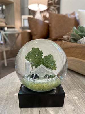 The FEARRINGTON SIGNATURE COLLECTION - FEARRINGTON BELTIE SNOWGLOBE from COOL SNOW GLOBES features a charming miniature rural scene complete with a white farmhouse, black cows, a tall silo, and green trees, all enclosed by a white picket fence. This delightful piece rests on a black square base against a white background.