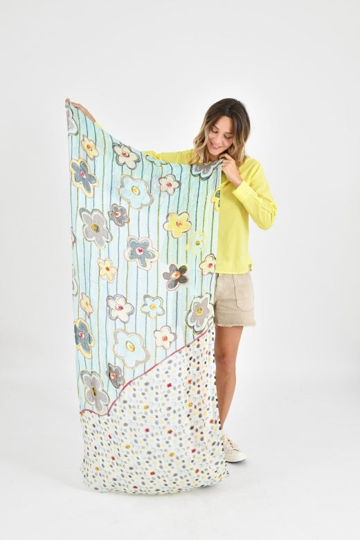 A woman with wavy brown hair wears a light yellow shirt and the STORIATIPIC - ANNE SCARF IN AQUA by STORIATIPIC, featuring colorful circular patterns. She smiles slightly at the camera, holding part of the hand-embroidered scarf.
