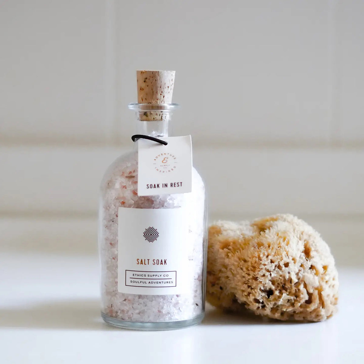 The ETHICS SUPPLY CO - SOAK IN REST BATH SALT SOAK, in a cork-sealed glass bottle and labeled "Salt Soak," sits beside a natural sea sponge. The setting features a white surface against a softly blurred neutral backdrop, with the subtle scent of gardenia filling the air.