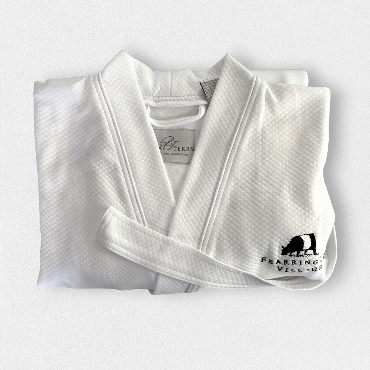 A neatly folded white kimono collar bathrobe adorned with a textured pattern. The bathrobe features an embroidered black bear and the text &quot;Fearrington Village&quot; on the bottom right side. Additionally, it boasts adjustable belt loops for a perfect fit. The label inside the collar reads &quot;F&quot;.