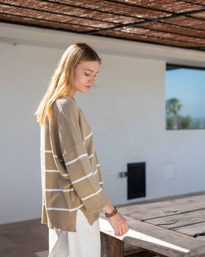 A woman wears the MERSEA - MONTAUK V-NECK SWEATER by MERSEA, a white oversized soft knit with blue and green striped collar, cuffs, and hem, ribbed v-neckline, styled over denim shorts and a light pink shirt.