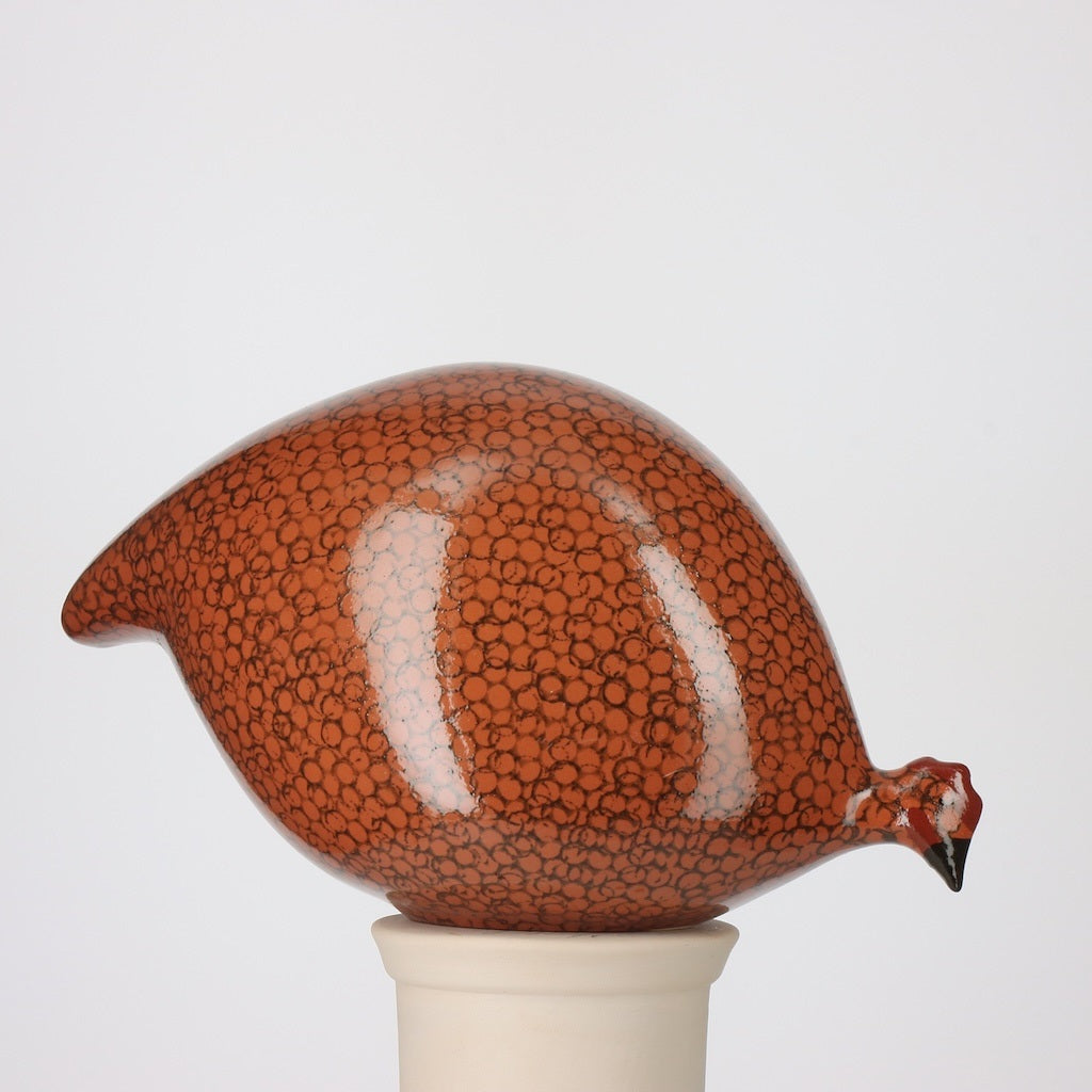 A ceramic sculpture from LES CERAMIQUES DE LUSSAN, the Pecking Ceramic French Guinea in Red Spotted Black, features a rounded bird form with small black spots on a reddish-brown surface, set on a white cylindrical base against a plain background—reminiscent of Heidi Caillard pottery and inspired by the iconic handmade ceramic French guinea hens.