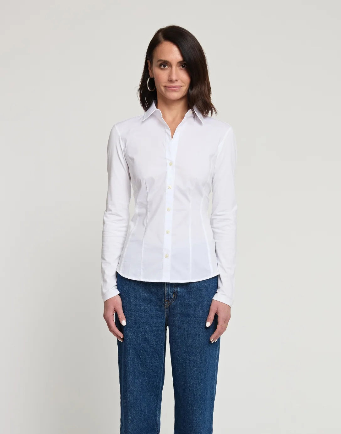 A woman with shoulder-length dark hair stands facing forward against a plain light background, wearing the HINSON WU Jamie Long Sleeve “T” Shirt in White crafted from stretch polished cotton and blue jeans, showcasing the refined finish of this versatile look.