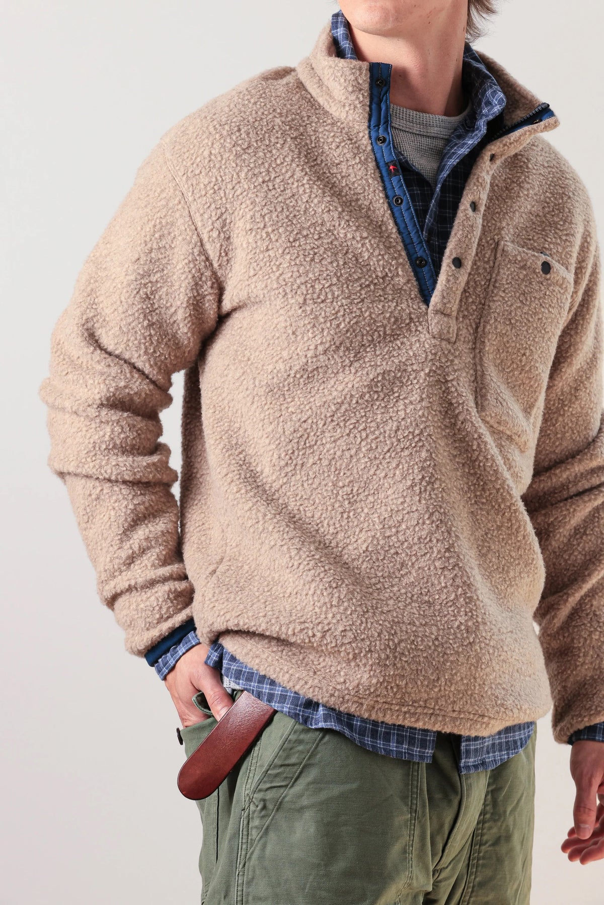 A person wearing the RELWEN Wooly Shag Snap-Zip in Dark Biscuit over a blue plaid shirt and green pants, with one hand in their pocket and a brown leather item partially visible at their waist.