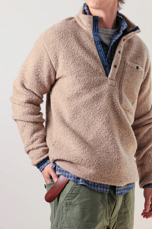 A person wearing the RELWEN Wooly Shag Snap-Zip in Dark Biscuit over a blue plaid shirt and green pants, with one hand in their pocket and a brown leather item partially visible at their waist.