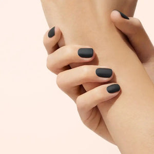 A hand with light blue painted nails holds a bottle of MANUCURIST - GREEN™ Nail Polish Matte Top Coat by MANUCURIST INC, featuring a black cap and clear plant-based formula, against a beige background.