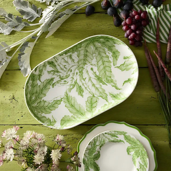 The CASKATA FREYA Coupe Oval Platter rests on a rustic green table amid carrots, grapes, greens, and white flowers. This porcelain piece features floral designs and is both dishwasher and microwave safe for easy care.