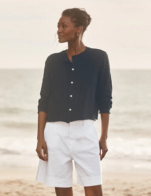 A woman stands on the beach, looking down and smiling as she wears the FRANK & EILEEN Jackie T-Shirt Cardigan in Vintage Jersey British Royal Navy, gently touching her collarbone with her left hand. The background is softly blurred with waves in view.