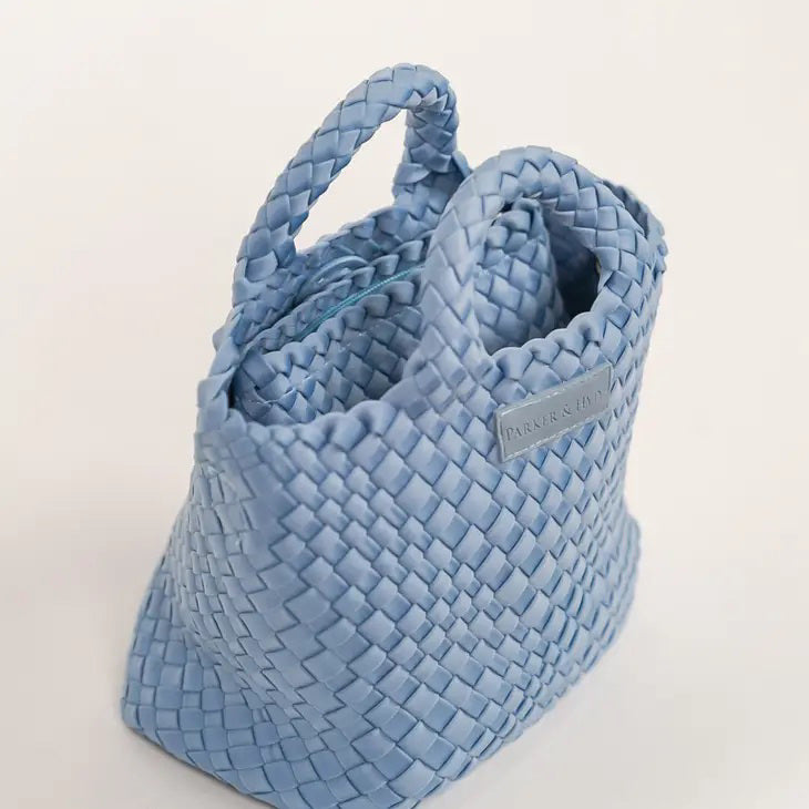 A woman with long brown hair, in a sleeveless cream knit top and blue jeans, holds a PARKER AND HYDE - MINI WOVEN TOTE BAG. The hand-woven tote by PARKER &amp; HYDE features a subtle checkered pattern with light blue accents.