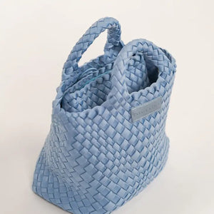 A woman with long brown hair, in a sleeveless cream knit top and blue jeans, holds a PARKER AND HYDE - MINI WOVEN TOTE BAG. The hand-woven tote by PARKER & HYDE features a subtle checkered pattern with light blue accents.