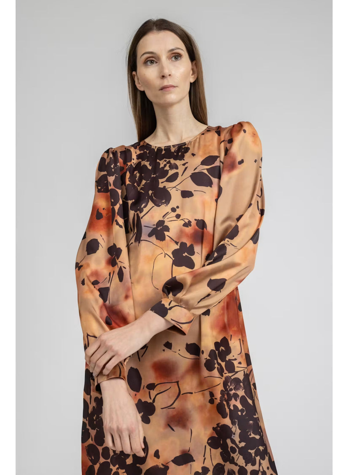 A woman stands with her back to the camera, wearing the MILVA MI - BURNT ORANGE PRINTED DRESS by MILVA—a long-sleeved, viscose midi dress in a rich brown shade accented with an orange and black botanical print—paired with black knee-high boots against a plain light background.