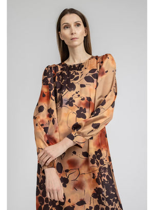 A woman stands with her back to the camera, wearing the MILVA MI - BURNT ORANGE PRINTED DRESS by MILVA—a long-sleeved, viscose midi dress in a rich brown shade accented with an orange and black botanical print—paired with black knee-high boots against a plain light background.