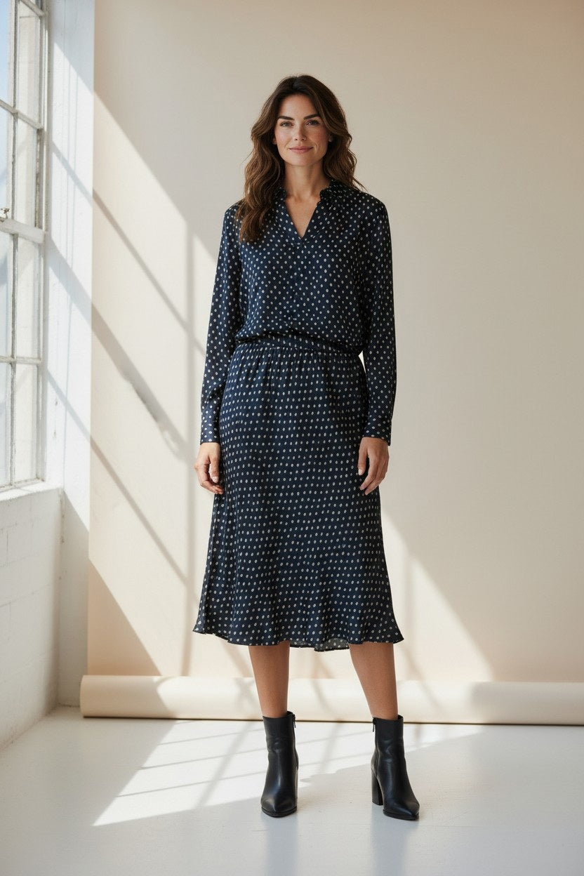 A woman stands in a bright room wearing the HARTFORD - CHORUS POPOVER SHIRT IN NAVY WITH WHITE DOTS, crafted by HARTFORD from soft printed viscose. She pairs it with black jeans and classic black loafers, one hand in her pocket as she gazes slightly to the side, radiating timeless French elegance. A small plant sits on a windowsill in the background.