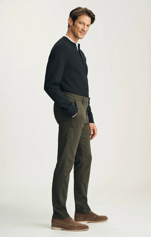 A person stands with their back to the camera against a neutral background, wearing the 34 HERITAGE - VERONA Straight Leg Chino Pant in Jasper High Flyer paired with a black long-sleeve shirt and brown suede shoes.