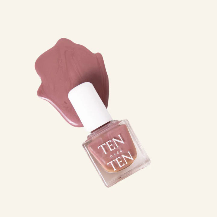 A glass bottle of TENOVERTEN - NAIL POLISH from the brand TEN OVER TEN, featuring a white cap and a dusty pink nail color. The front label displays "TEN OVER TEN" in white uppercase letters. This vegan, eight-free formula ensures both beauty and peace of mind. The background is plain and light-colored.