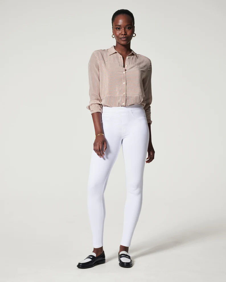 A woman stands in a studio against a neutral background wearing the SPANX JEANNISH ANKLE LEGGING, styled with a light brown striped blouse, black-and-white loafers, minimal jewelry, and high-waisted tummy shaping fit.
