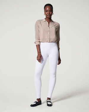 A woman stands in a studio against a neutral background wearing the SPANX JEANNISH ANKLE LEGGING, styled with a light brown striped blouse, black-and-white loafers, minimal jewelry, and high-waisted tummy shaping fit.